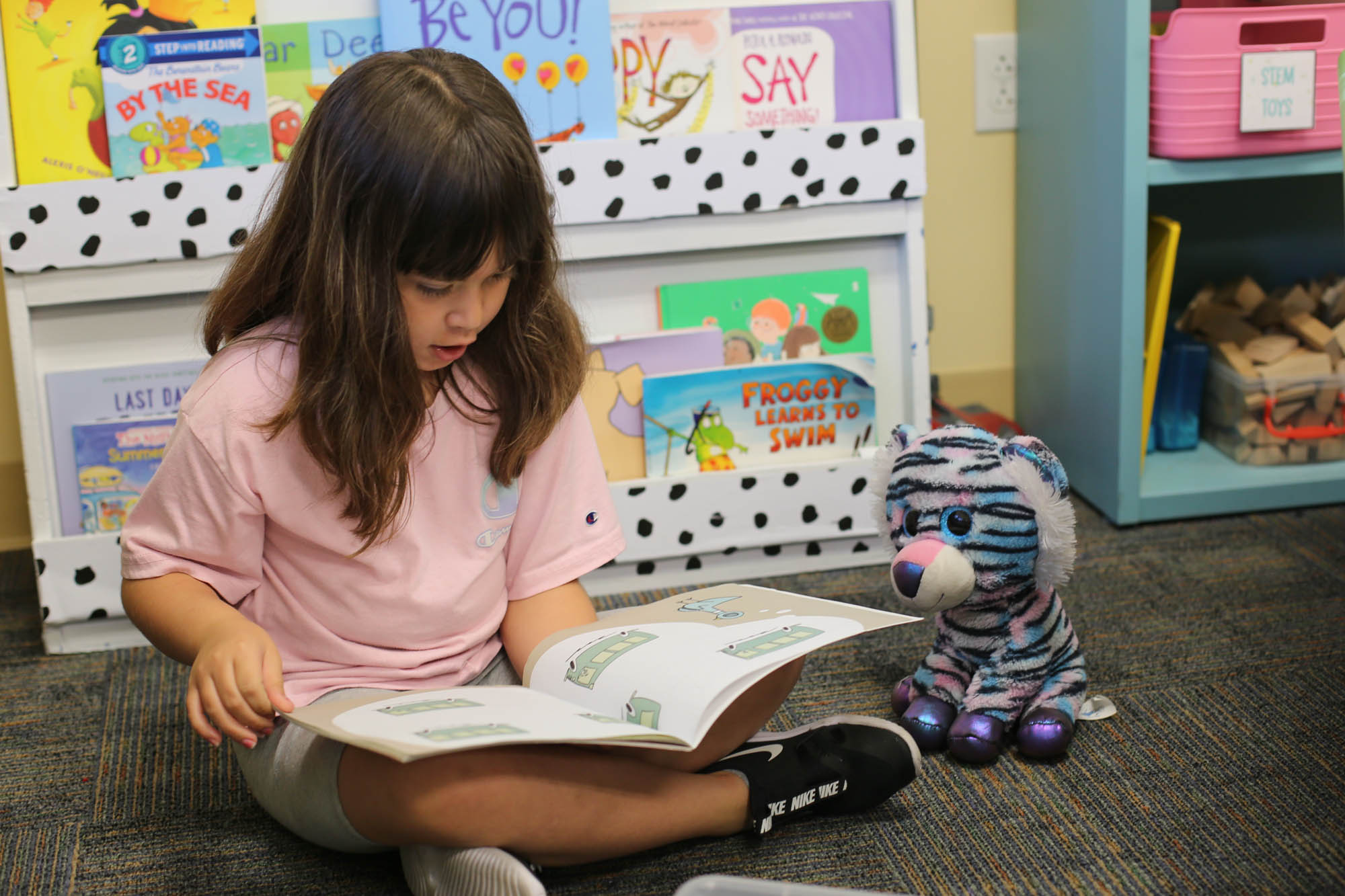 A young girl in a pink shirt reads on the floor of the classroom in front of a bookcase