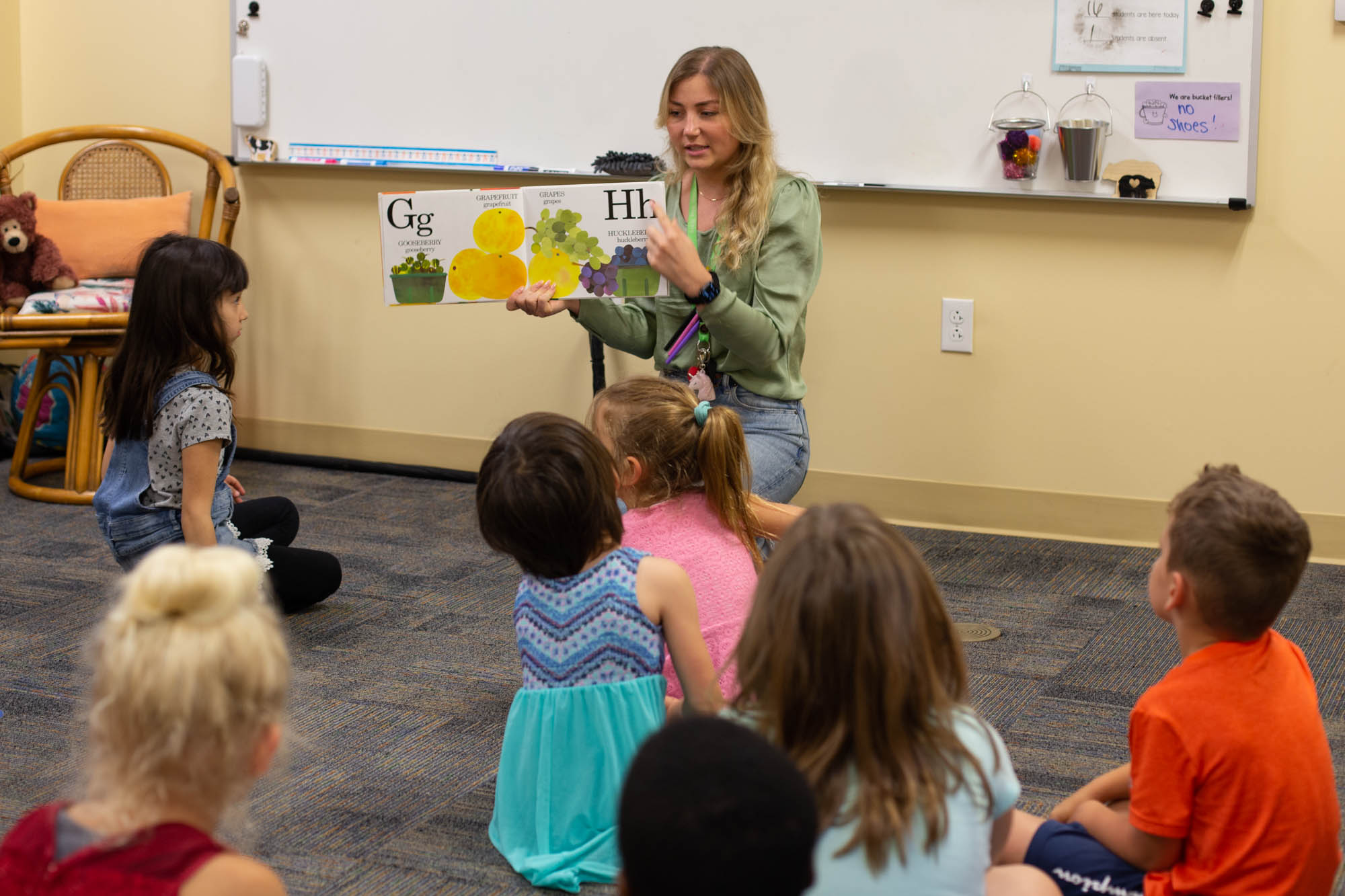 A teacher is kneeled on the floor with her early learning students reading them a book about the alphabet.