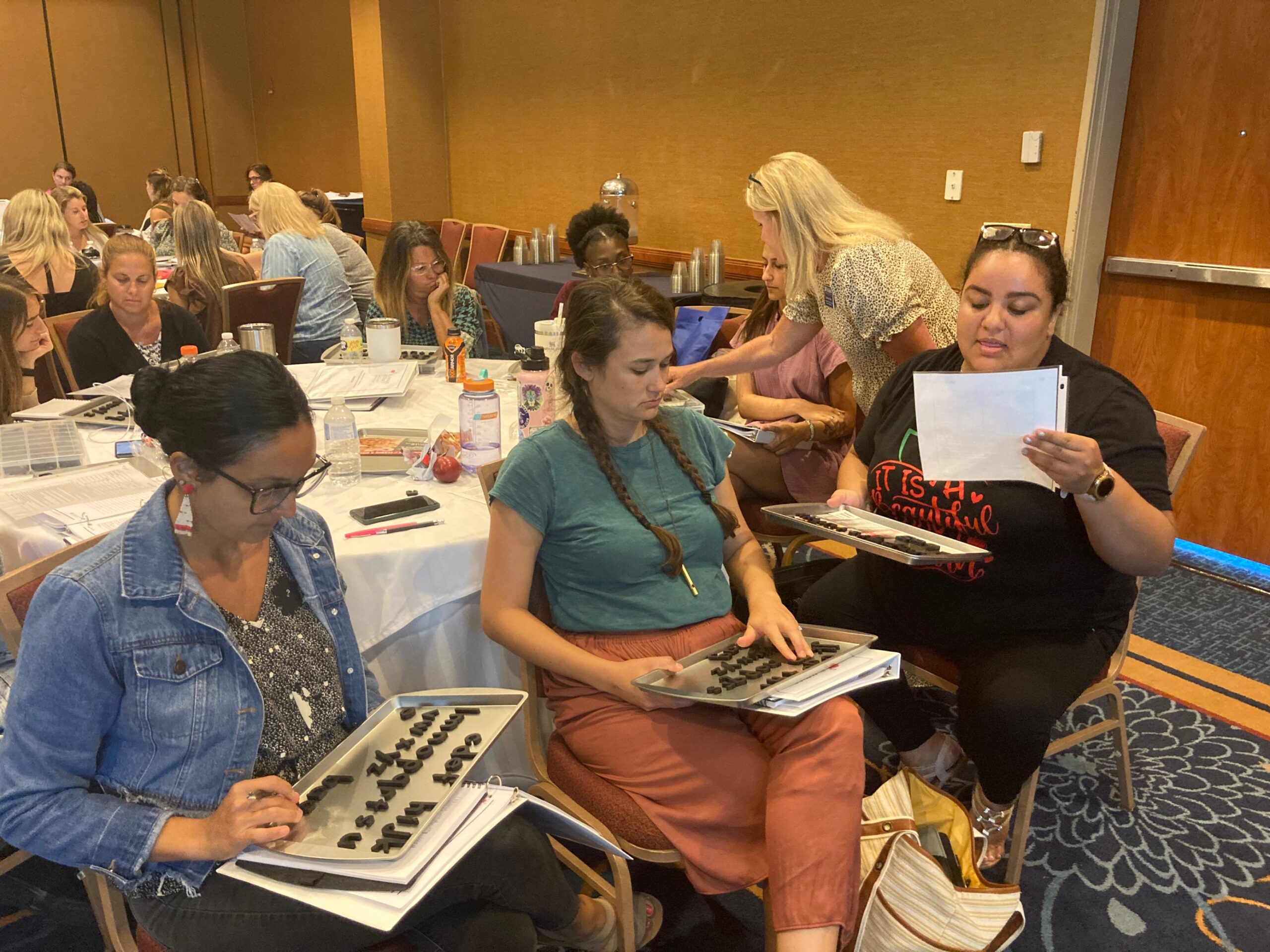Three teachers sit together at a round table and practice with manipulative letters - a teaching practice to help developing readers learn common sounds and word patterns.