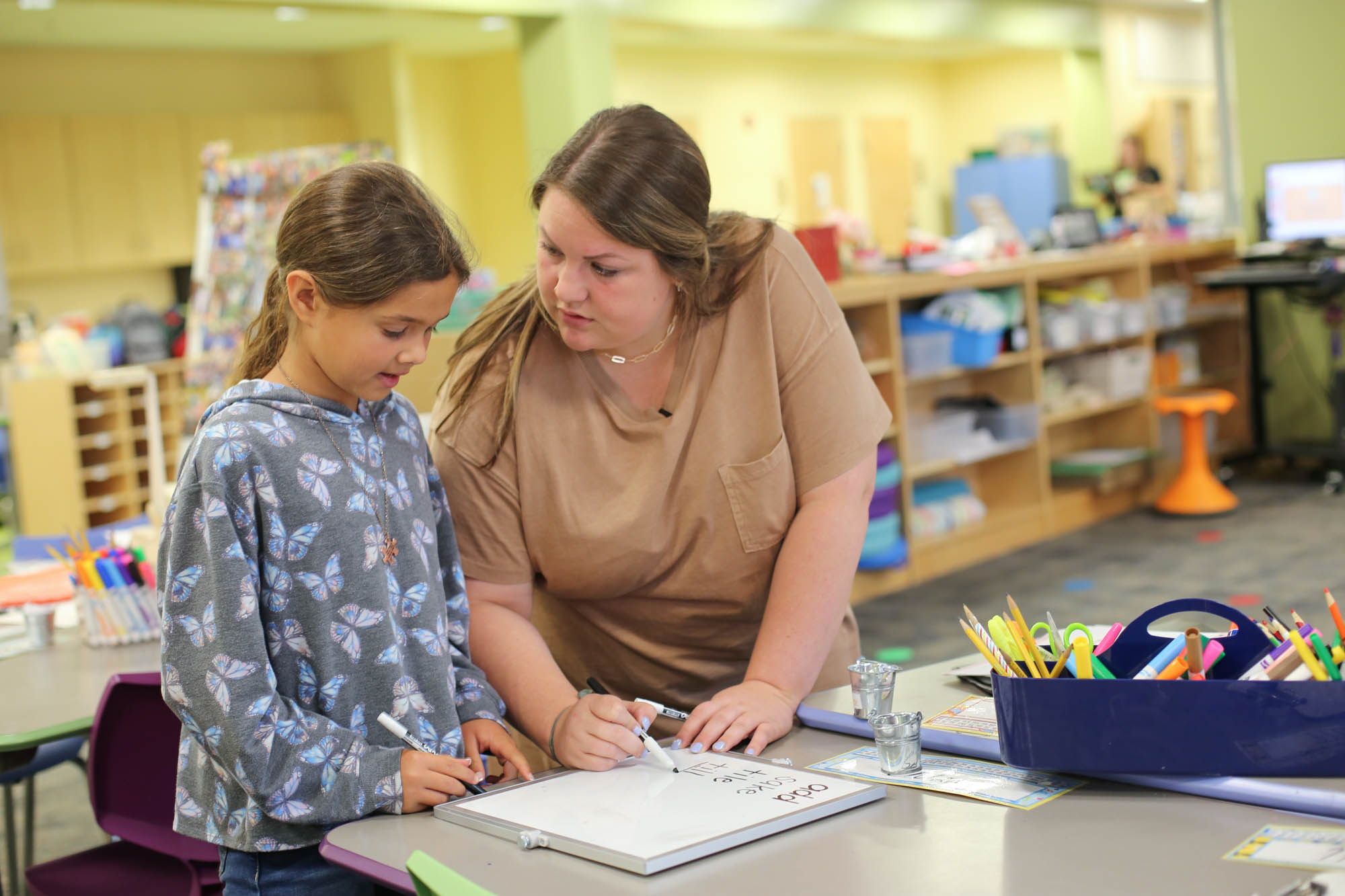 A teacher and student stand together in a classroom practicing spelling on a white board.