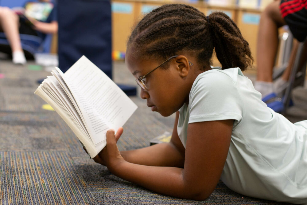 A young elementary student lays on the floor of the classroom reading a book.