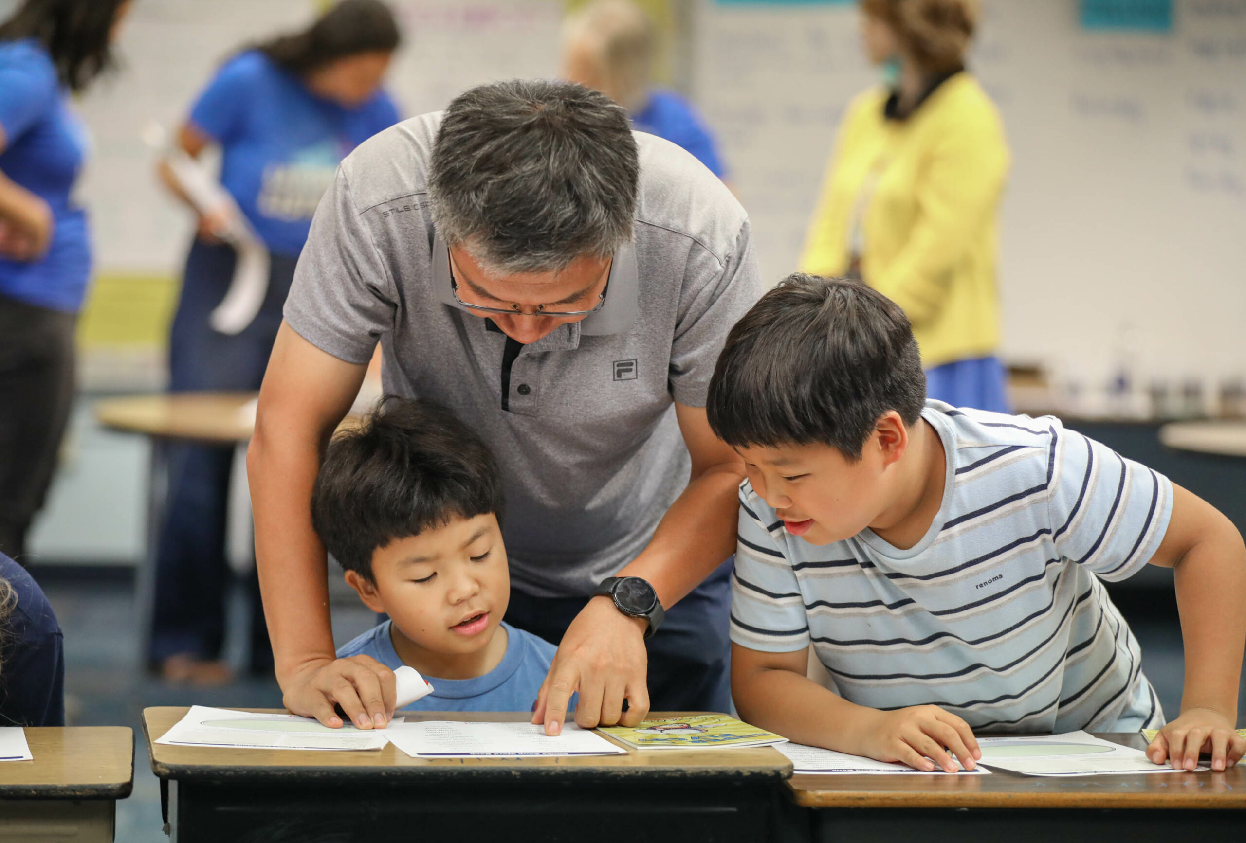 A father leans over a desk to help his two sons work on an activity together.