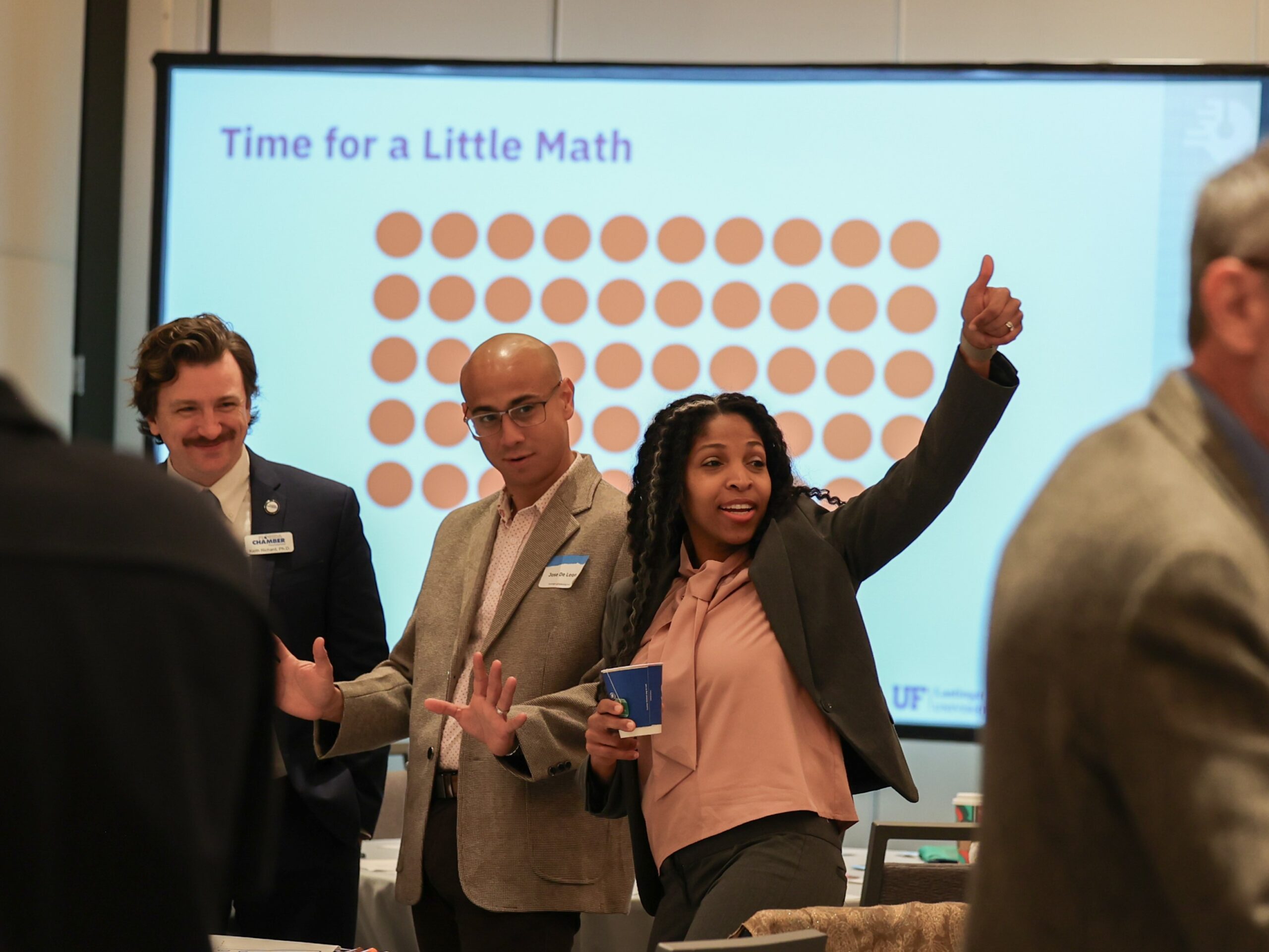 A photo of three Math Summit attendees standing in front of a Presentation that says, "Time for a Little Math." Two men are smiling and looking off to the side, and a woman next to them giving a thumbs up. 