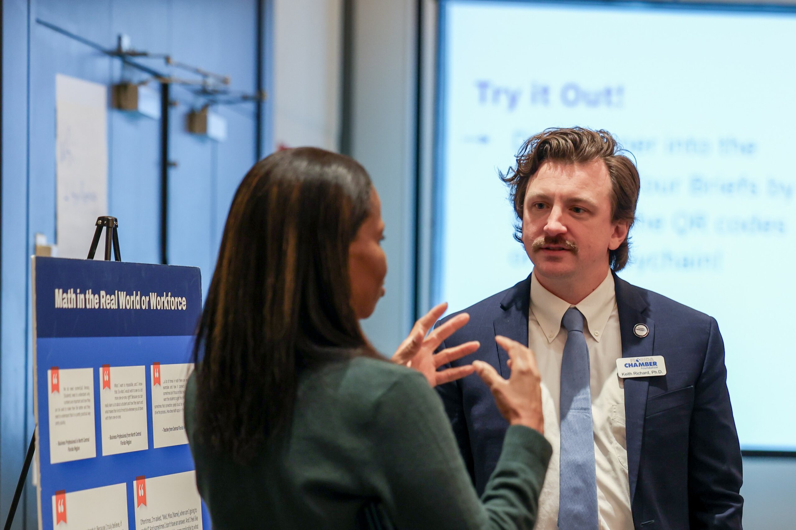 A photo of two Math Summit attendees discussing the presentation board they're standing next to, titled "Math in the Real World or Workforce."